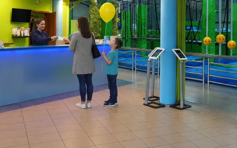 Mama z synem trzymającym balon odbiera kartę przyjęcia przy recepcji Jumpout (obraz wygenerowany przez AI)