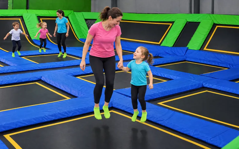 Mama w różowej koszulce skacze za rękę z kilkuletnią córką na trampolinie w hali Jumpout, w tle limonkowe ściany i inne dzieci (obraz wygenerowany na tle prawdziwej hali Jumpout)