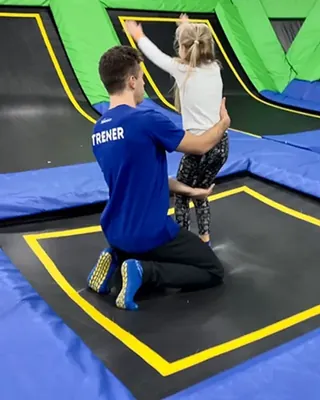 Trener w koszulce z napisem TRENER klęczy na trampolinie i asekuruje skok dziewczynki z rękami w górze