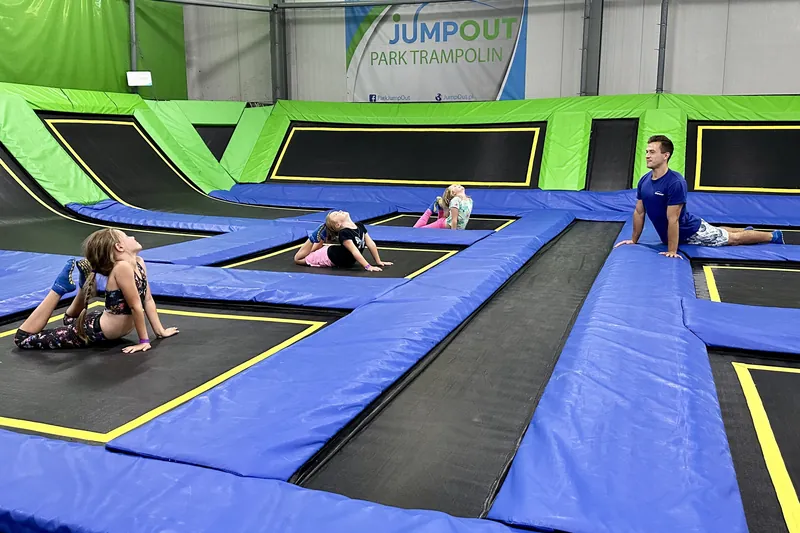 Trzy dziewczynki i trener rozciągają się na trampolinach w Jumpout, w tle baner Jumpout Park Trampolin