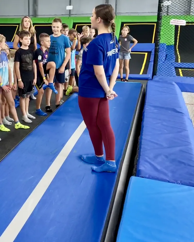 Trenerka w niebieskiej koszulce z napisem TRENER stoi na ścieżce airtrack przed szeregiem dzieci w skarpetach antypoślizgowych, w tle pola trampolin Jumpout