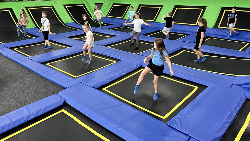 Hala Jumpout w głąb: dzieci skaczą na czarnych polach trampolin z żółtym obrysem, między polami niebieskie maty, w tle limonkowe ściany skośne