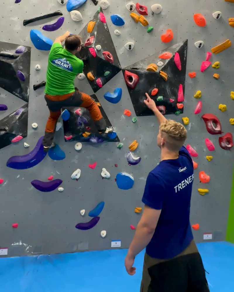 Trener w koszulce z napisem TRENER wskazuje chwyt wspinaczowi na ściance boulderowej Jumpout