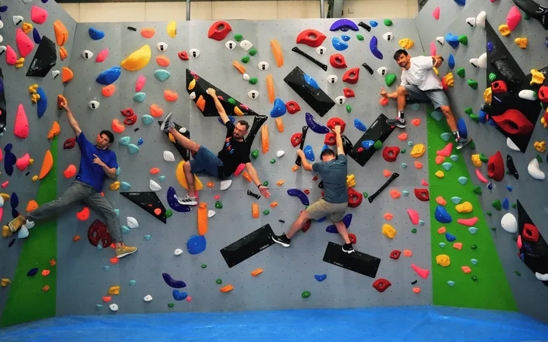Czterech wspinaczy na trasach szerokiej ścianki boulderowej z kolorowymi chwytami, pod ścianą niebieski materac