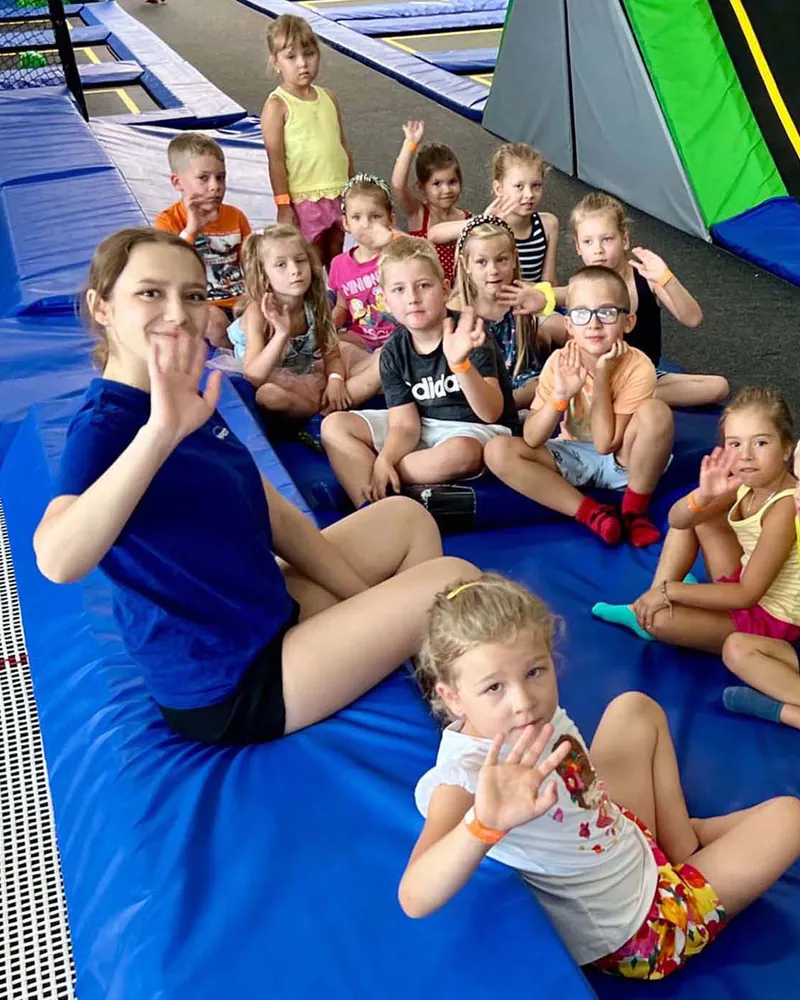 Animatorka w granatowej koszulce siedzi z grupą urodzinową na miękkiej macie przy trampolinach, wszyscy machają na powitanie przed zabawą