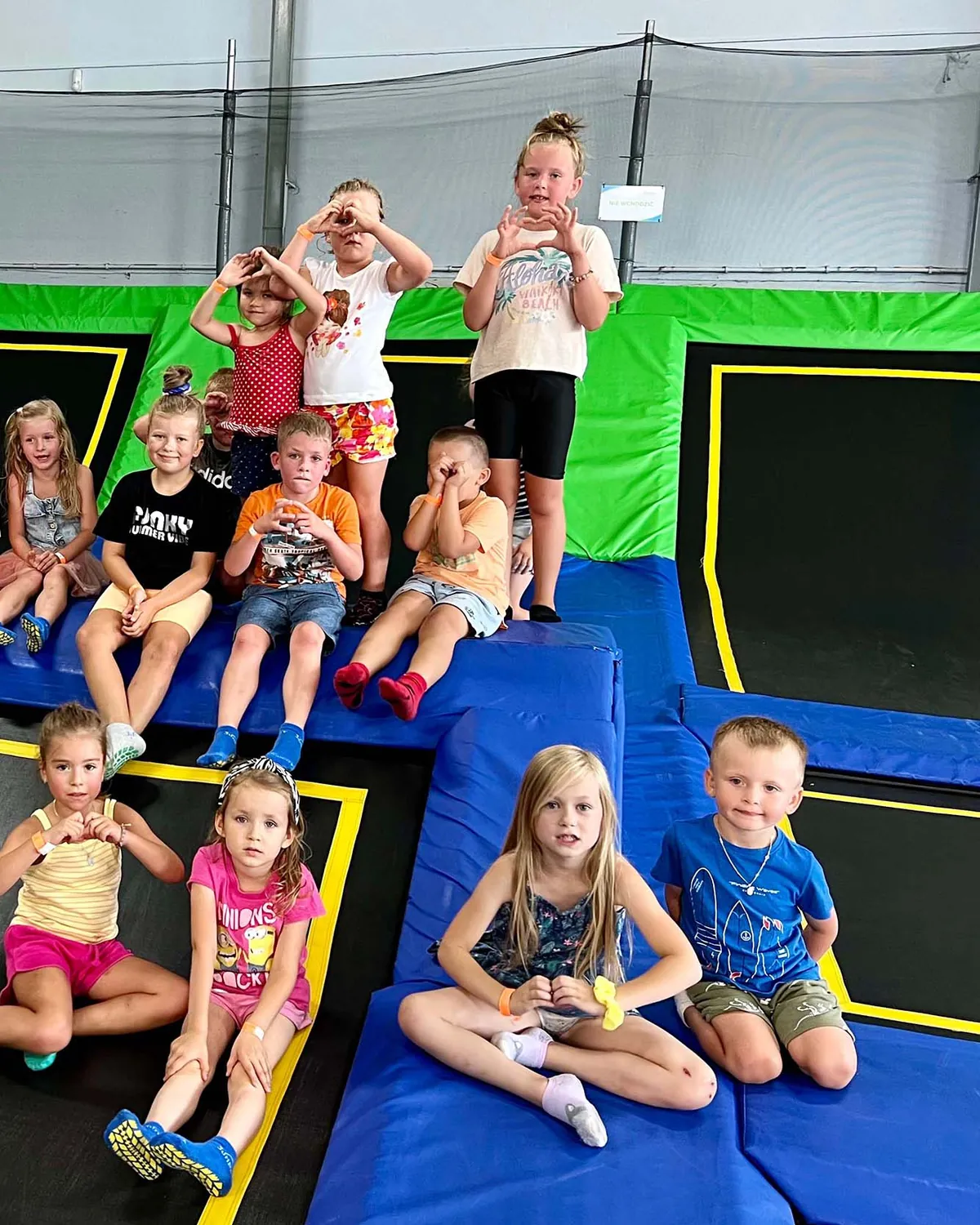 Grupa urodzinowa siedzi na polach i materacach trampolin, dzieci pokazują serduszka z dłoni